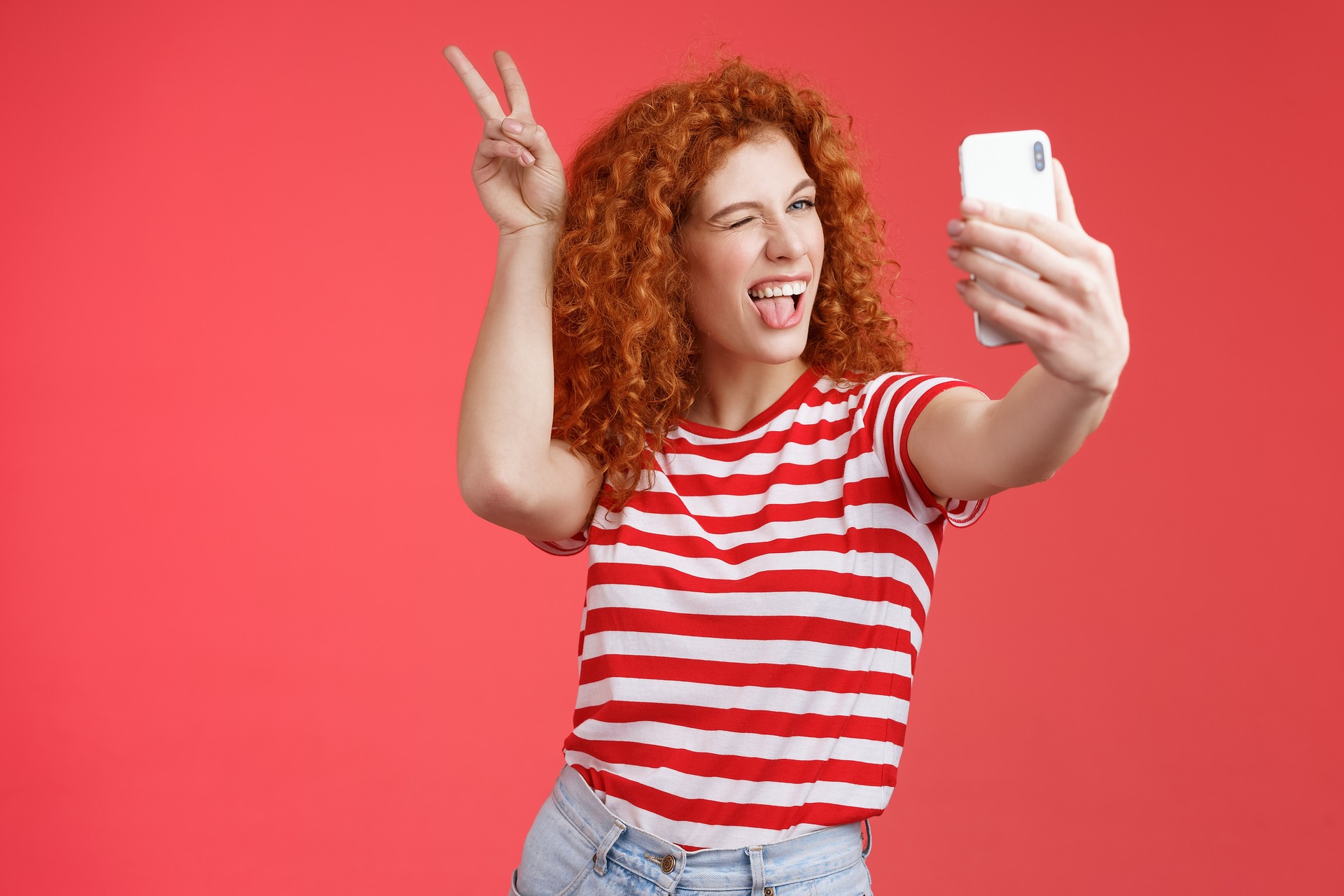 Photo of a white woman with red curly hair, wearing a red and white horizontal striped shirt and jeans in front of a red background, taking a selfie of herself while winking, sticking out her tongue, and giving herself bunny ears by mahbubhasan2550 on Pixabay