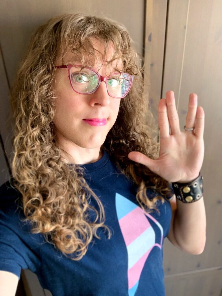 Me in a dark blue t-shirt with the Star Trek delta on it, in trans pride flag colors. I’m making the live long and prosper Vulcan salute with my hand.