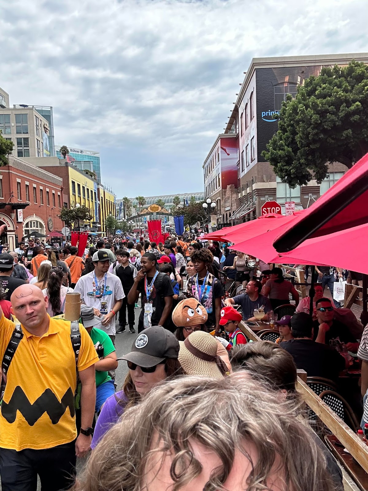 A shot of a long line and a very large crowd of nerds on a cute street, next to diners on a restaurant patio, with my curly bangs in the foreground