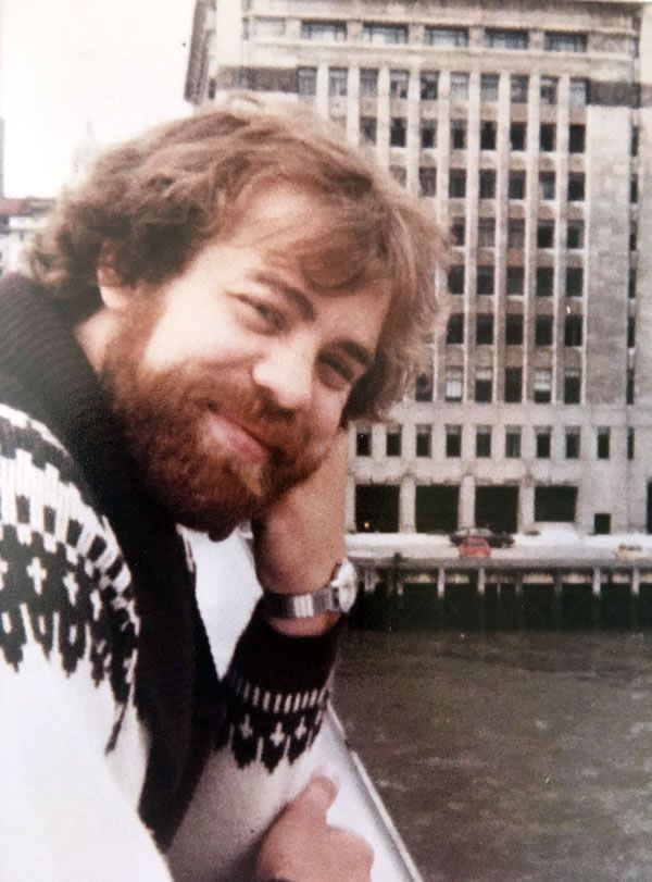 A photo of my dad, with short wavy brown hair and a brown beard, leaning on the railing of a bridge over a river, looking at the camera and smiling.