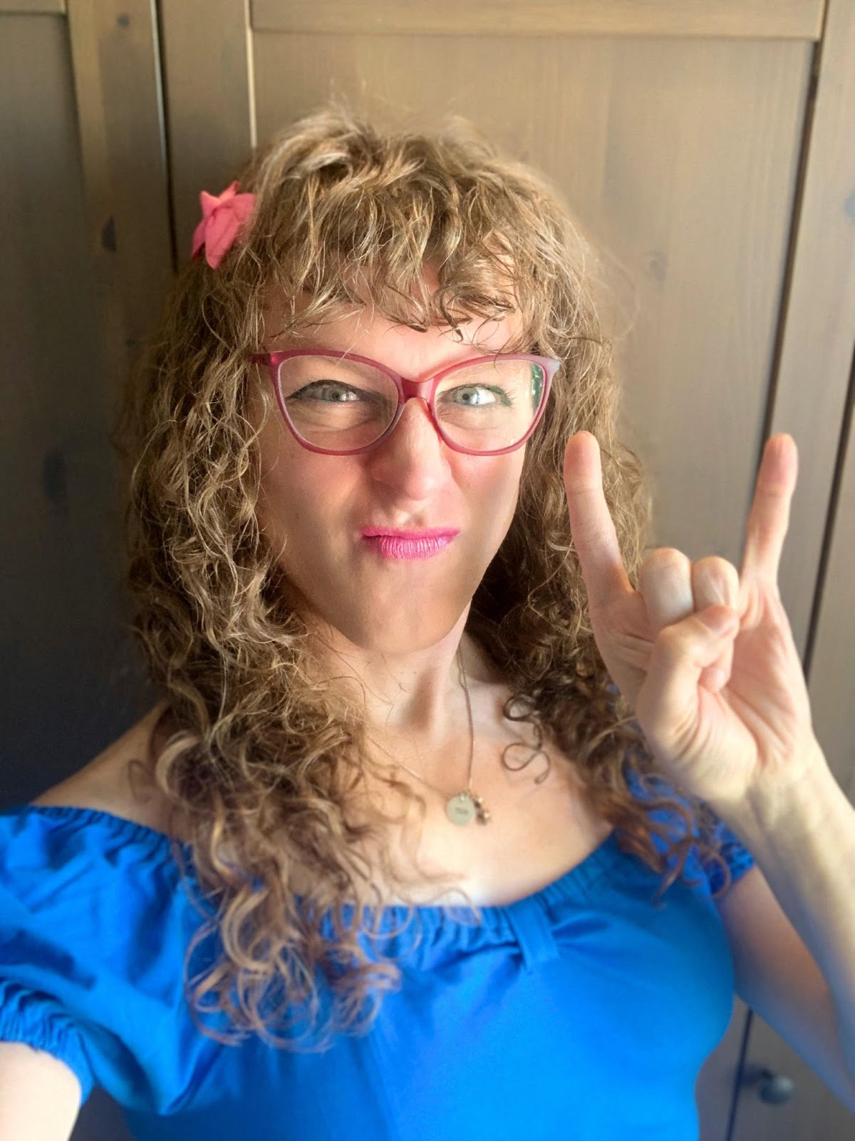 me with long curly brown hair and curly bangs, with a pink bow in my hair, in dark eyeliner and light pink lipstick, wearing pink-framed cat-eye glasses and a blue off-the-shoulder top... and I'm throwin' up the horns!