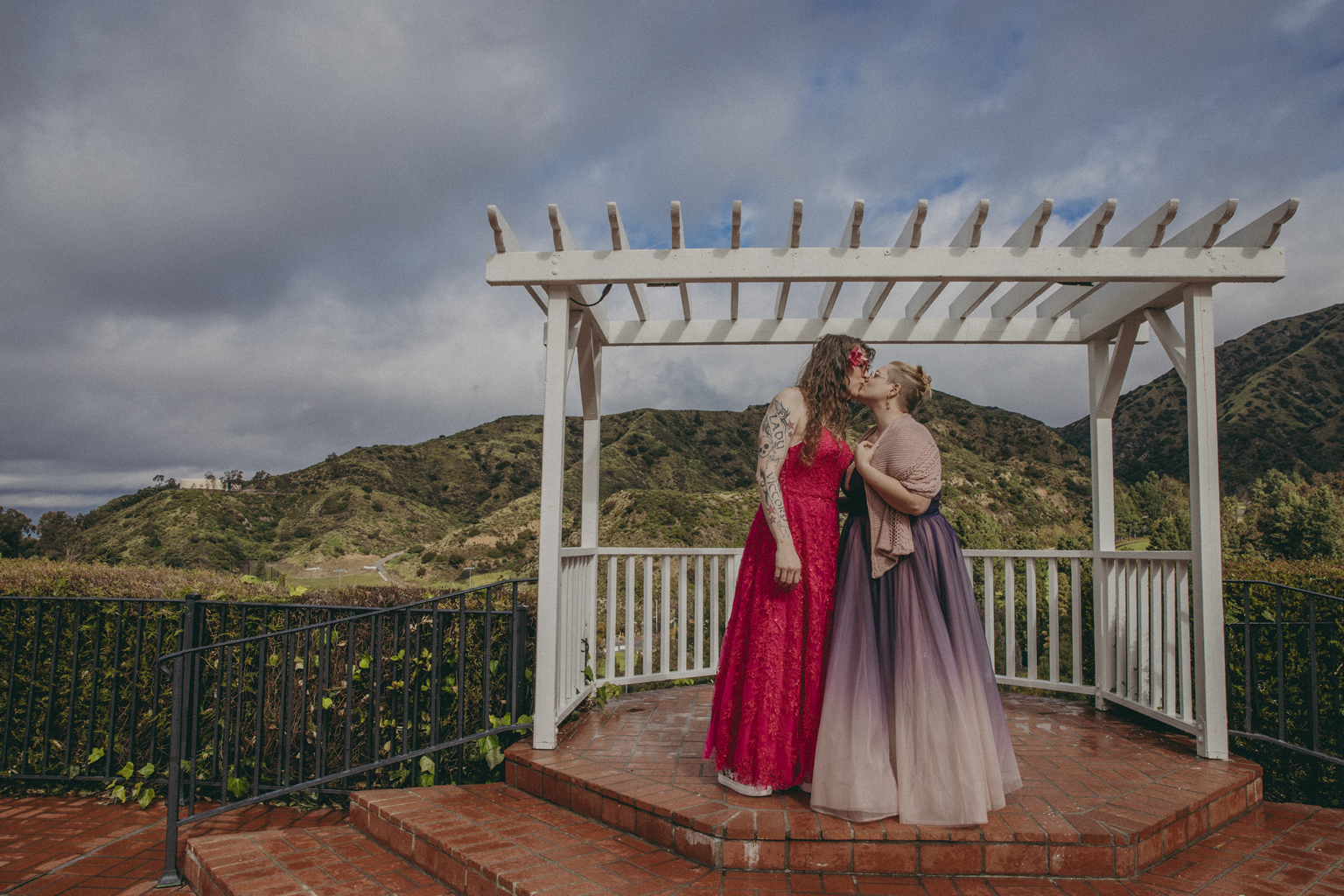 (Photo by Kim Newmoney) Me in my fuchsia dress, kissing my wife Susan in a purple dress, under a white lattice arch, in front of green mountains and under a very blue but very cloudy sky. The ground is wet.