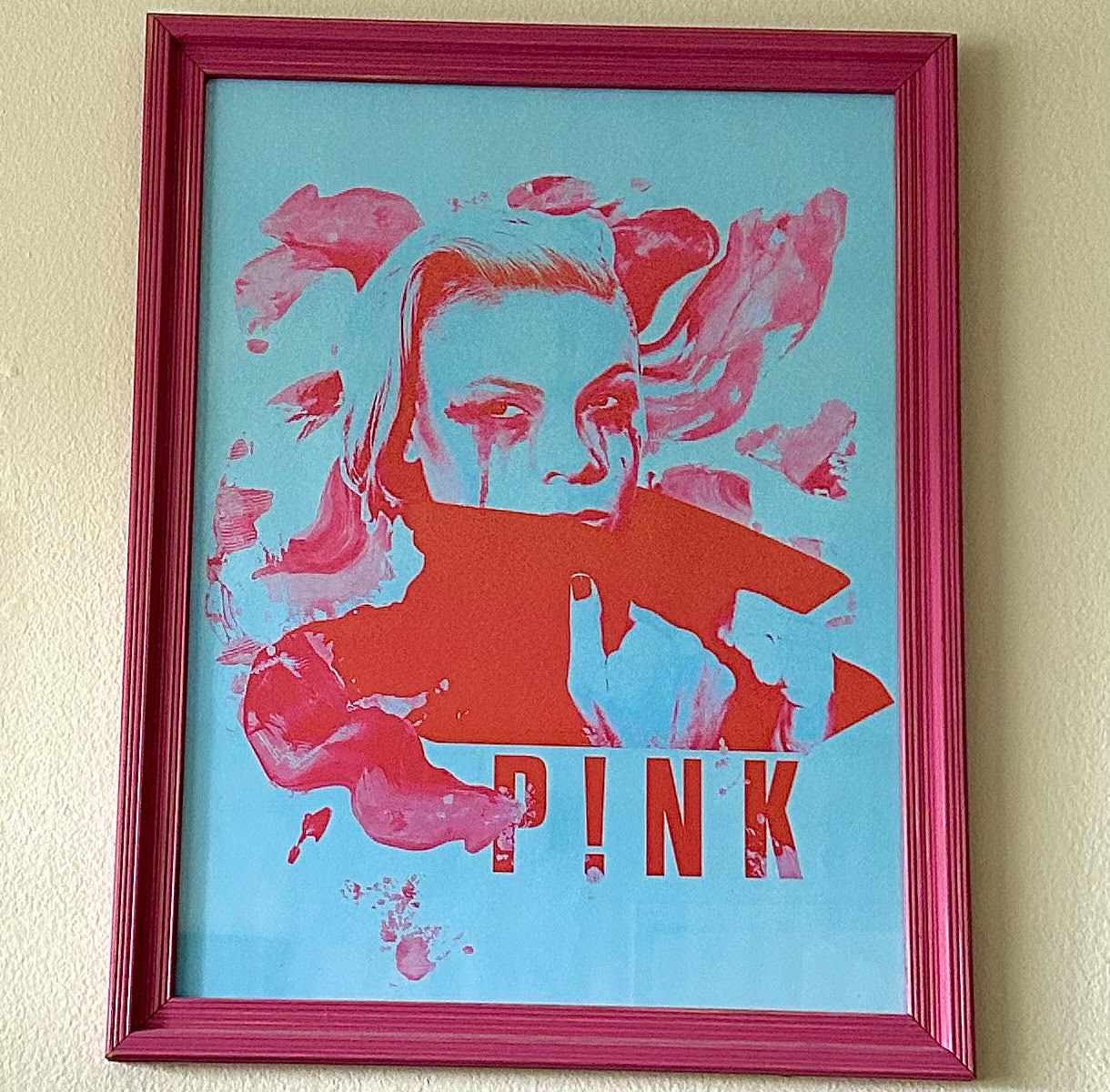 A light blue poster with pink print on it, showing a photo of P!nk in a coat with a large collar, and splotches of pink splattered around. Her hair is swept to her right side, and her eyeliner and mascara is running down from her eyes. The poster is under glass inside a pink frame.