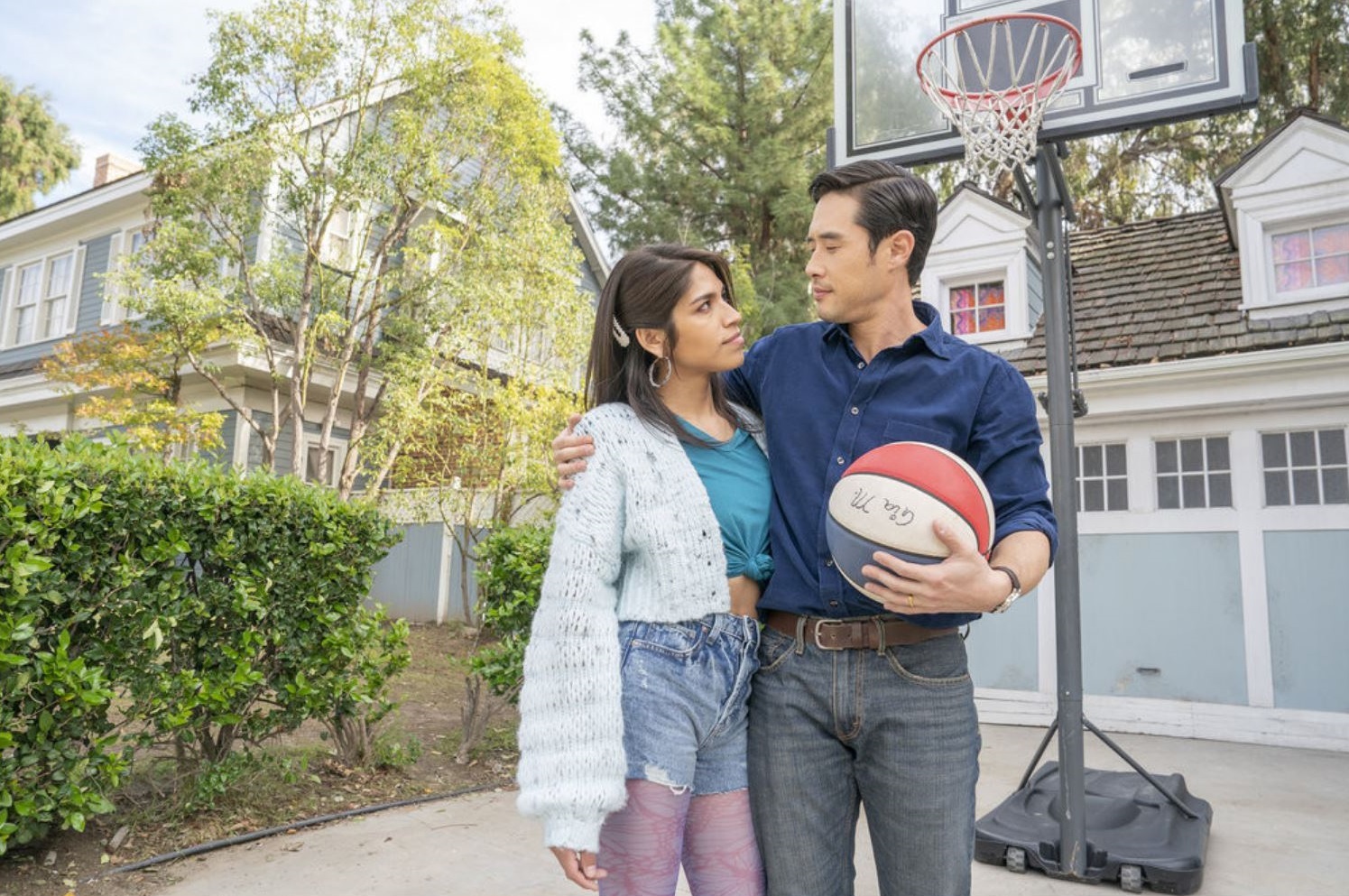 Raymond Lee, as Ben Song, holding a basketball with one hand and his other hugging Josielyn Aguilera as Gia Mendéz. They're standing on a driveway in front of a house with a basketball hoop behind them.