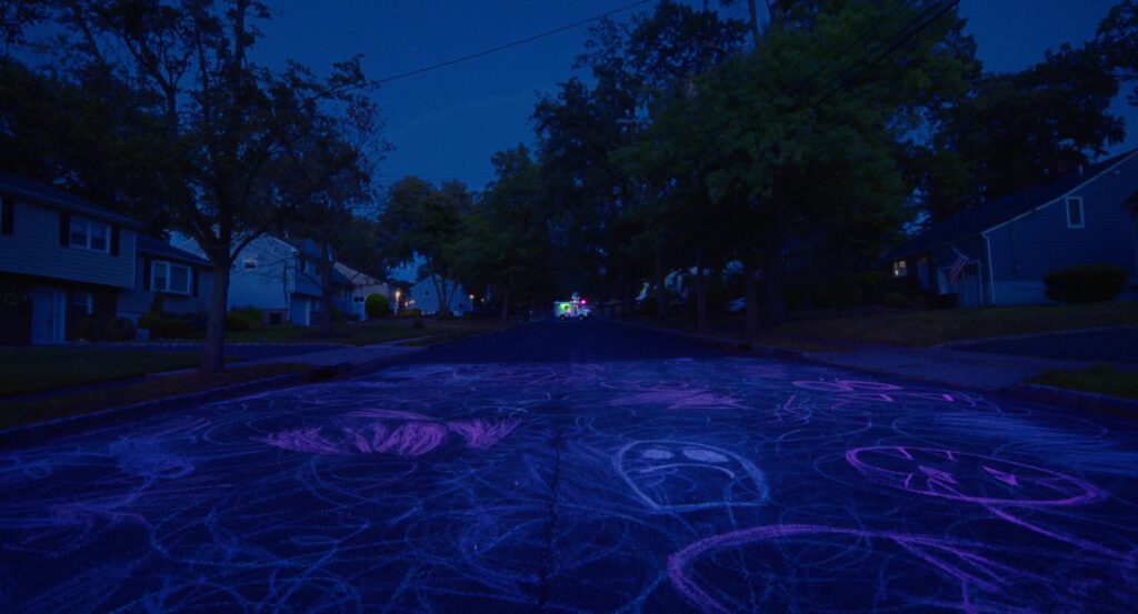 A nighttime shot of a street in a neighborhood, houses on either side. The street is covered in a jumble of pink and blue chalk scribbles and drawings, most prominent is a sad blue ghost. At the end of the street, very small, is an ice cream truck lit up in green and pink.