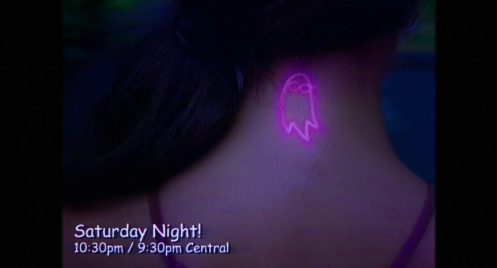 The back of Isabel’s neck with the lit up neon-pink ghost-in-glasses-with-open-eyes tattoo, and the text “Saturday Night! 10:30pm / 9:30pm Central”.