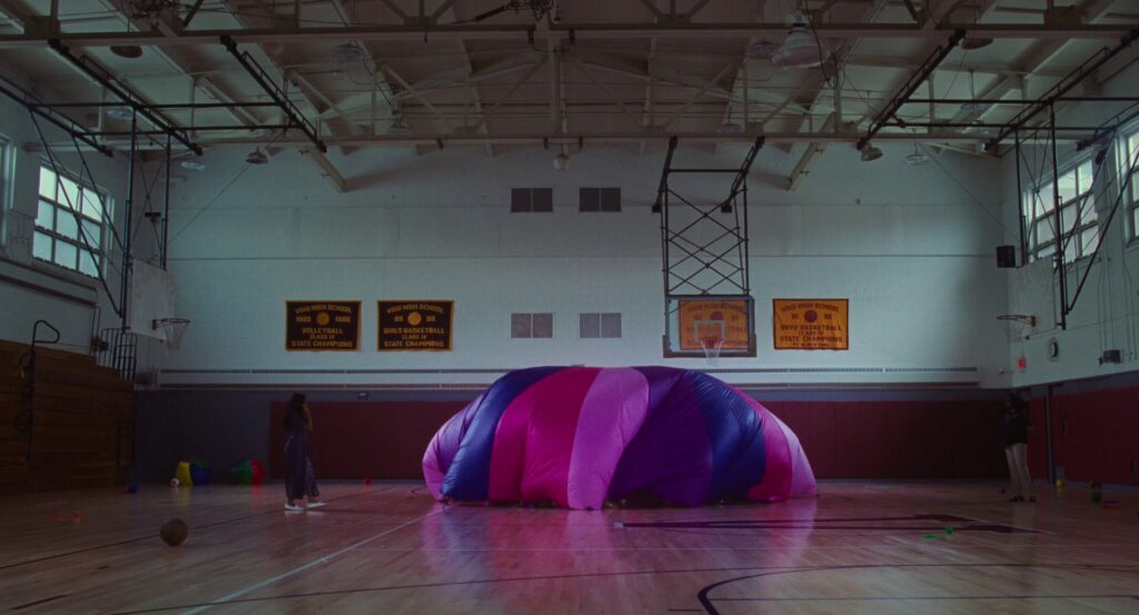 A pink, white, blue, and purple parachute with air trapped under it to turn it into a floaty dome inside a school gymnasium