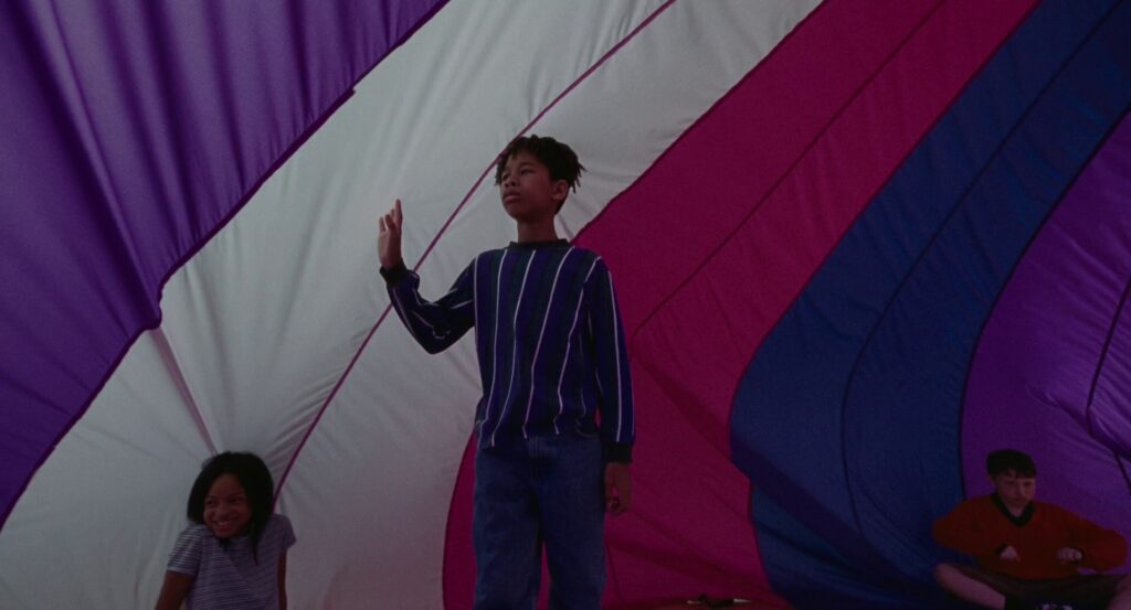 Young Owen walking inside the purple, white, pink, and blue parachute in his school gymnasium.