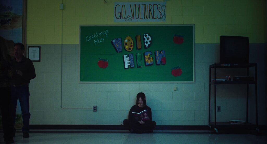 Kinda goth-Maddy sits under a sign that says “Go Vultures” above a blackboard that says “Greetings from Void High.”