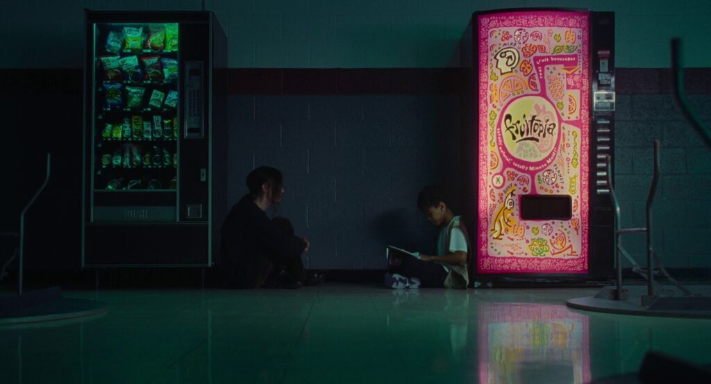 Maddy and Owen sit between two vending machines, Maddy with her back to a green one full of snacks, and Owen with his back to a pink Fruitopia one.