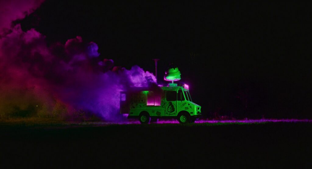 A closer shot of the far-off ice cream truck from the opening shot, the truck lit in only green, with pink smoke rising from behind it.