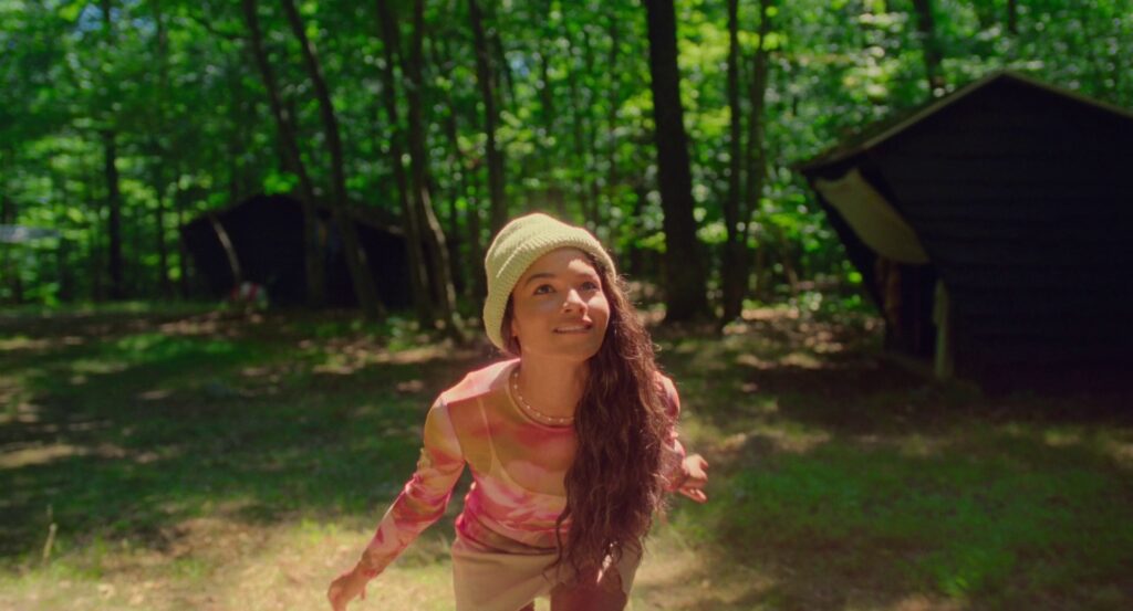 Isabel, in a pink top, runs out of a green forest with a smile on her face, eyes looking up.