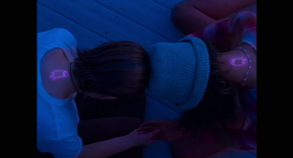 
Tara and Maddie sitting cross-legged, with their heads bent and tops of their heads touching, showing matching pink ghost tattoos on the backs of their necks.