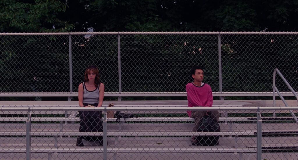 Maddy and Owen sitting on bleachers at the school football field. Owen is in a pink sweatshirt and Maddy is in a long skirt and femme tank-top, with a much more femme presentation 