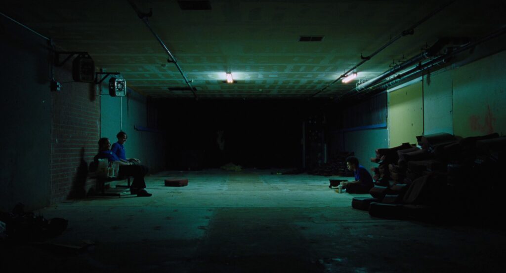 Owen sits across from his manager and his manader’s toadie in the big empty… warehouse? The lighting is very green, there’s a yellow wall behind Owen, a red wall behind the manager, and everyone’s wearing blue shirts.