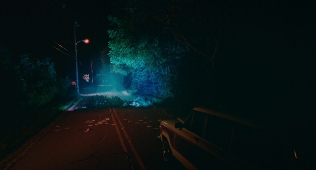 Owen’s car stopped in the road as a downed power line is in front of him. The lighting is red and yellow where his car is, and blue/green by the power line. A street light further back appears pink.