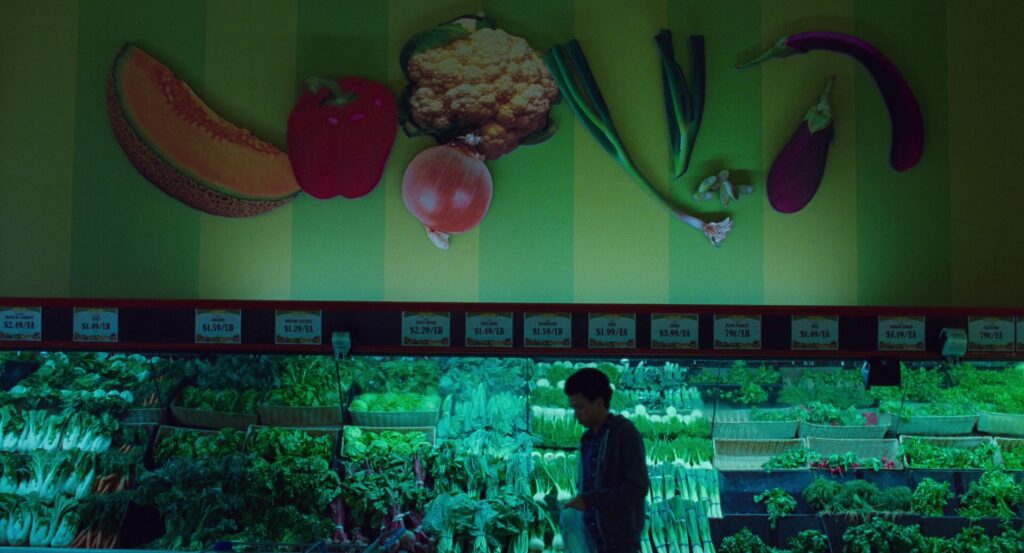 Owen walking in front of a wall of produce at a grocery store, it’s all lit in green and large images of various produce on are the wall above his head