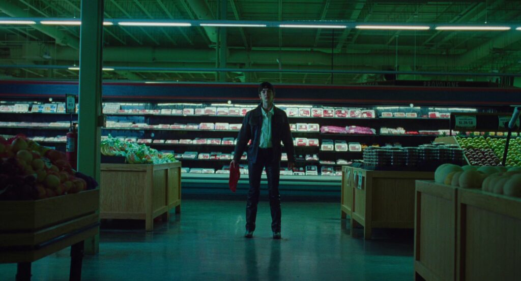 Maddy stands in the middle of the grocery store, a refrigerated meat section behind her. She holds a raw steak in one hand, and is dressed very gender-neutral.