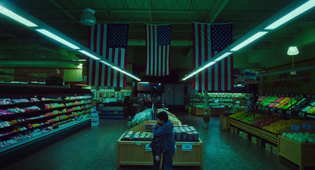 Owen hugs Maddy in the middle of the grocery store, bathed in green light and under three American flags. 