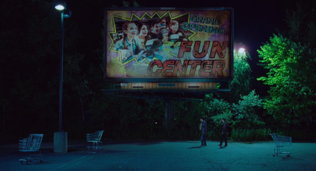 Owen and Maddy walk beneath a billboard for the grand opening of the Fun Center, surrounded by empty shopping carts.