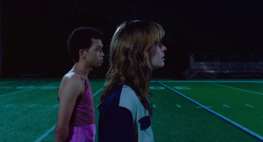 Owen, in his pink dress, walks with Maddy on a green football field.