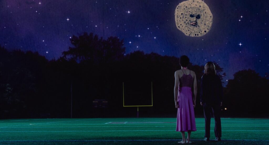 Owen, in his pink dress, stands next to Maddy on the green football field, looking up at a blue sky full of stars and Mr. Melancholy’s face sticking out his tongue in the moon.