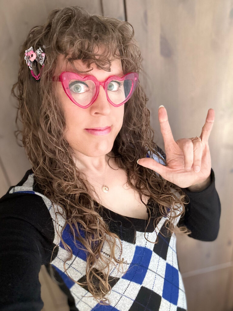 Me in a blue, gray, and black argyle sweater with pink heart-shaped glasses and a pink skull bow in my long brown curly hair, making the "i love you" american sign language sign