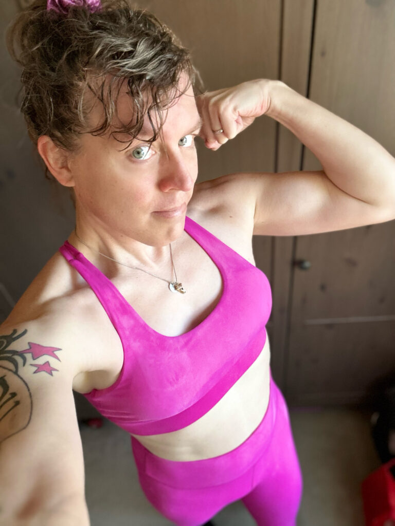 Me in a hot pink sports bra and tights, flexing my left bicep which looks barely defined at all, shot from a higher angle looking down.