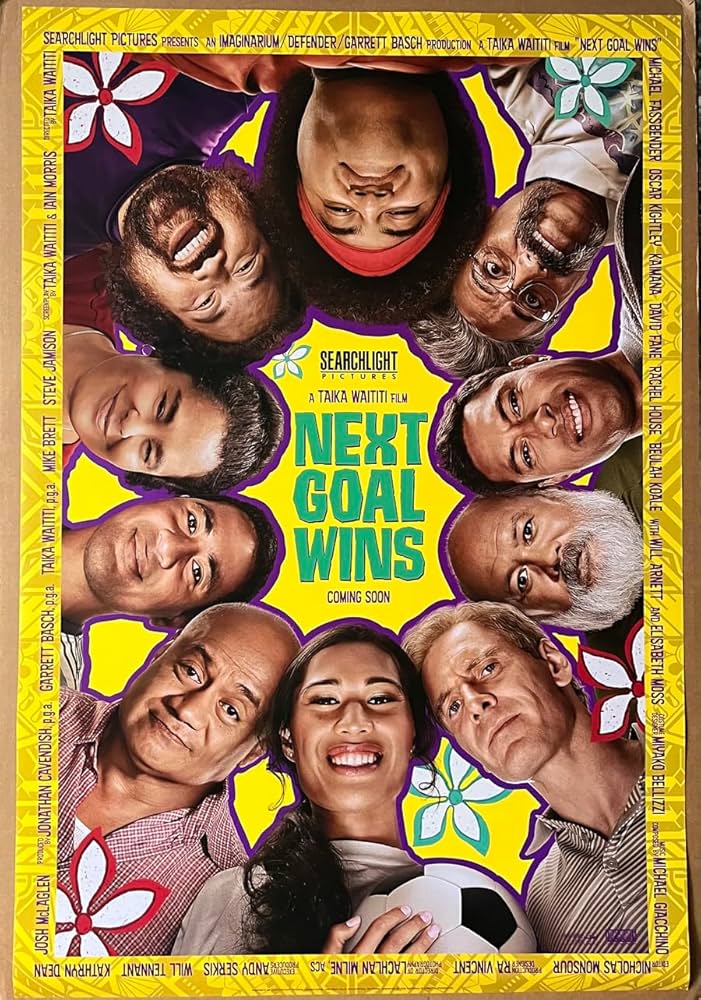 Next Goal Wins promo poster looking up, as if from the ground, toward the cast which are in a circle around the poster, as if in a huddle, looking down. Jaiyah holds a soccer ball.